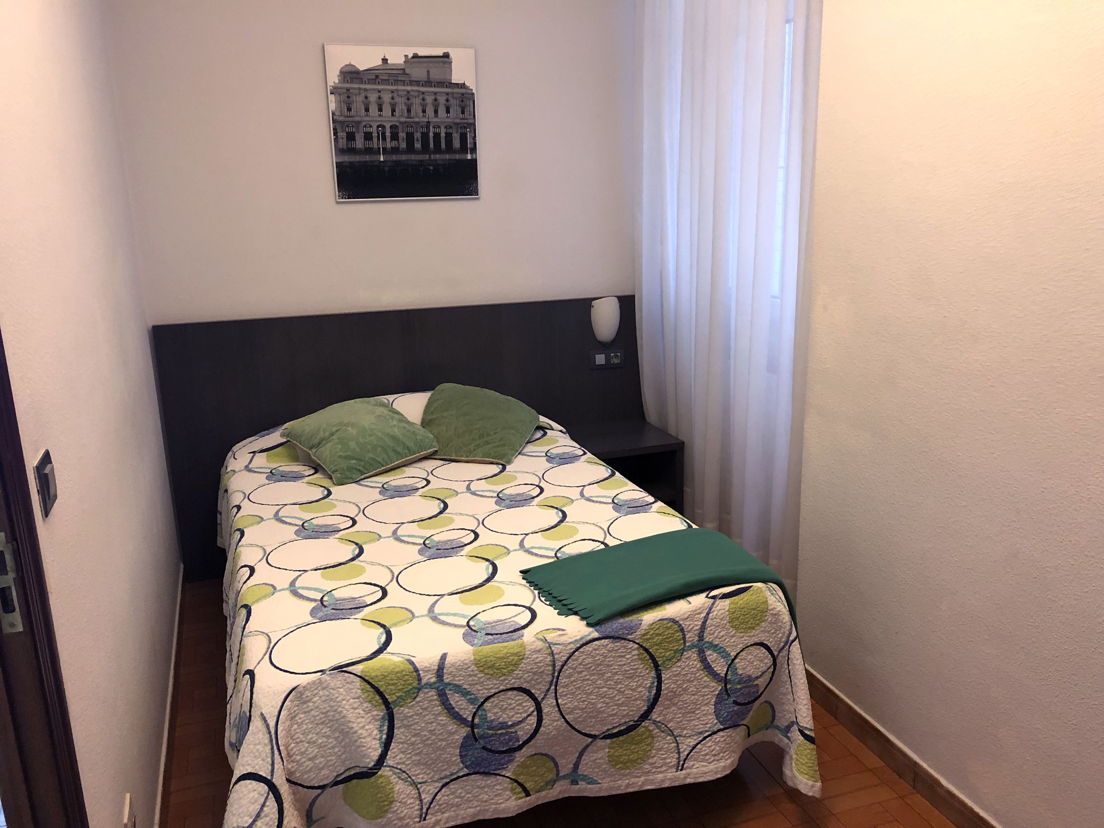 Een klein, netjes opgemaakt bed met een beddengoed met patroon en twee groene kussens, een donker hoofdbord, een klein nachtkastje en een foto van een gebouw aan de muur.
