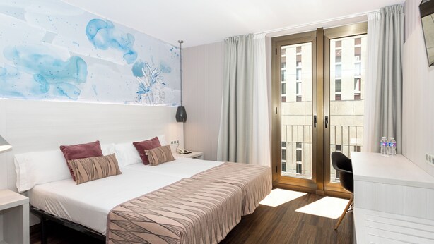 Habitación de hotel con cama, mesita de noche, escritorio, silla y vista al exterior a través de una puerta.