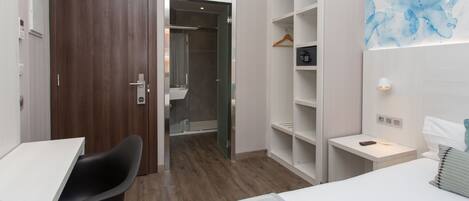 A hotel room with a wooden door, a desk with a chair, a bed, a walk-in closet, and a bathroom visible through an open door.