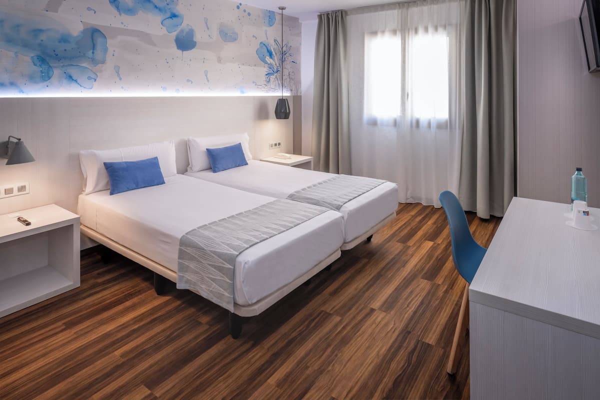 Une chambre d’hôtel avec deux lits, un bureau, une chaise et une grande fenêtre avec des rideaux.