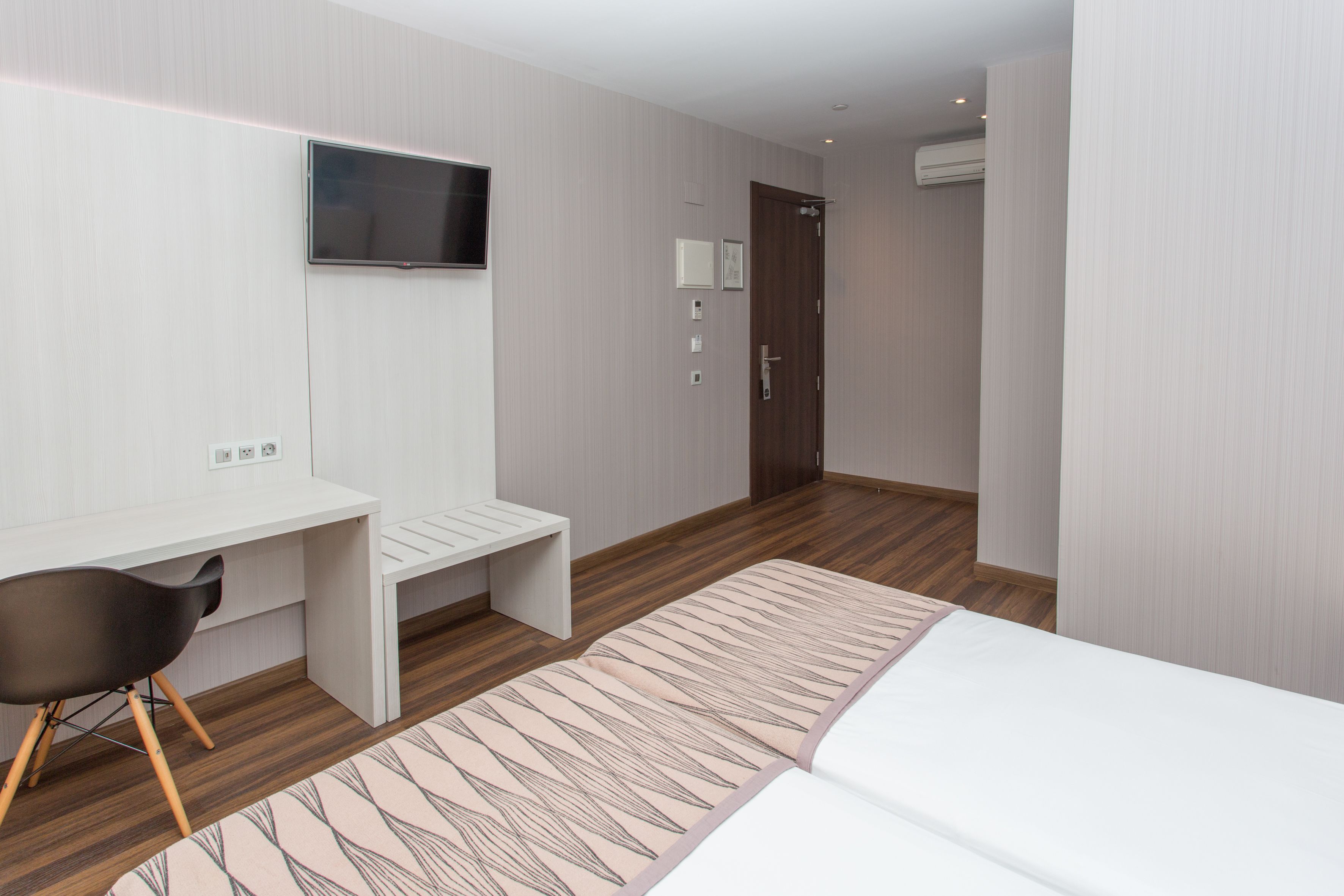 A modern hotel room with a bed, a desk, a chair, a TV, and a door.