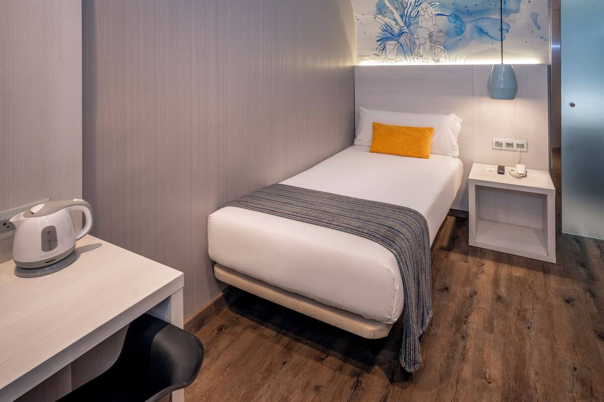 Une chambre d’hôtel comprenant un lit, un bureau avec une bouilloire, une table de chevet et un mur orné d’un tableau abstrait bleu.