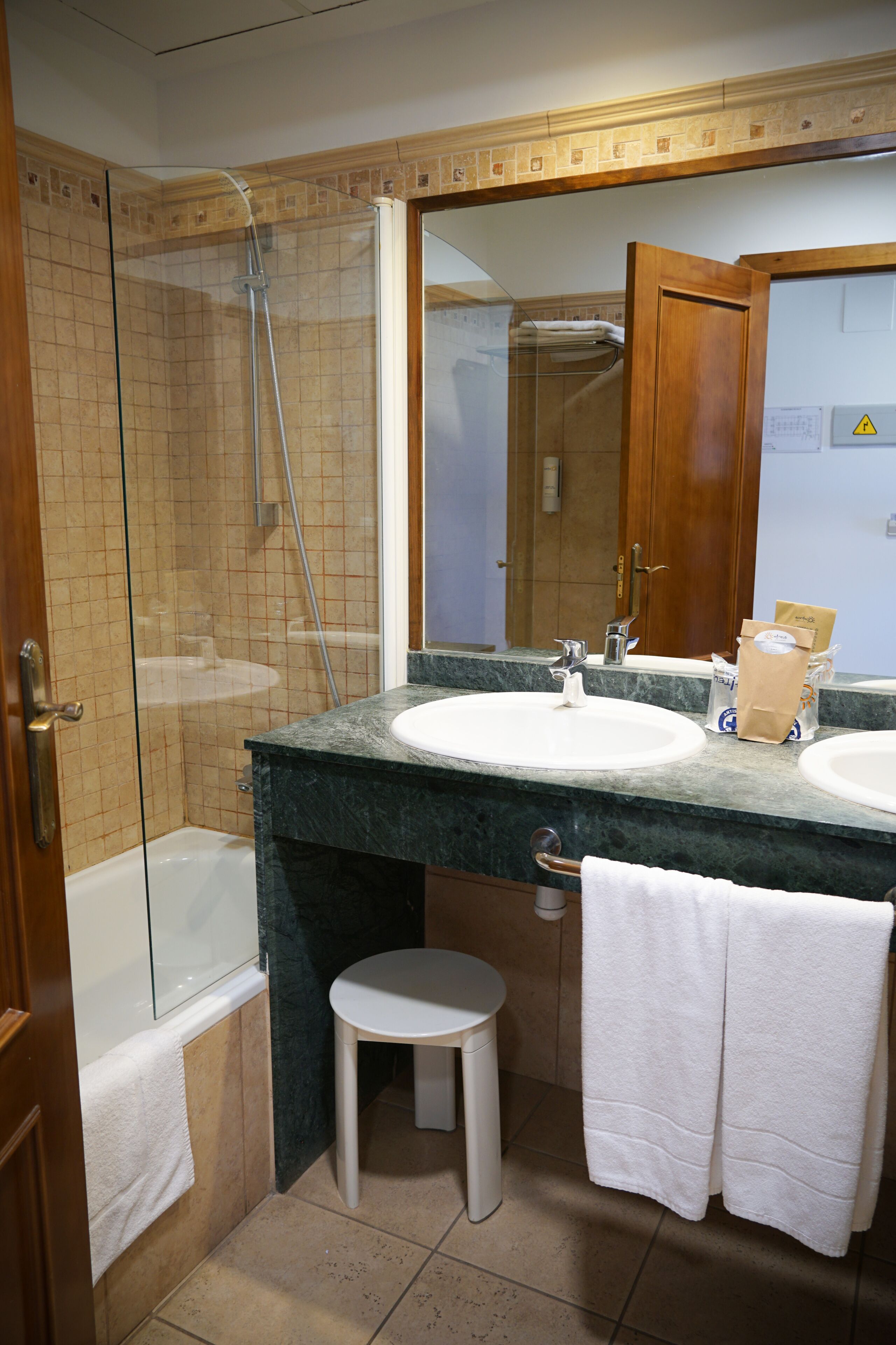 Un baño con ducha de vidrio, dos lavamanos, un espejo y un taburete pequeño.