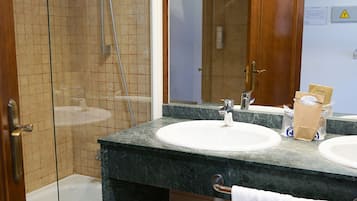 Un baño con ducha de vidrio, dos lavamanos, un espejo y un taburete pequeño.