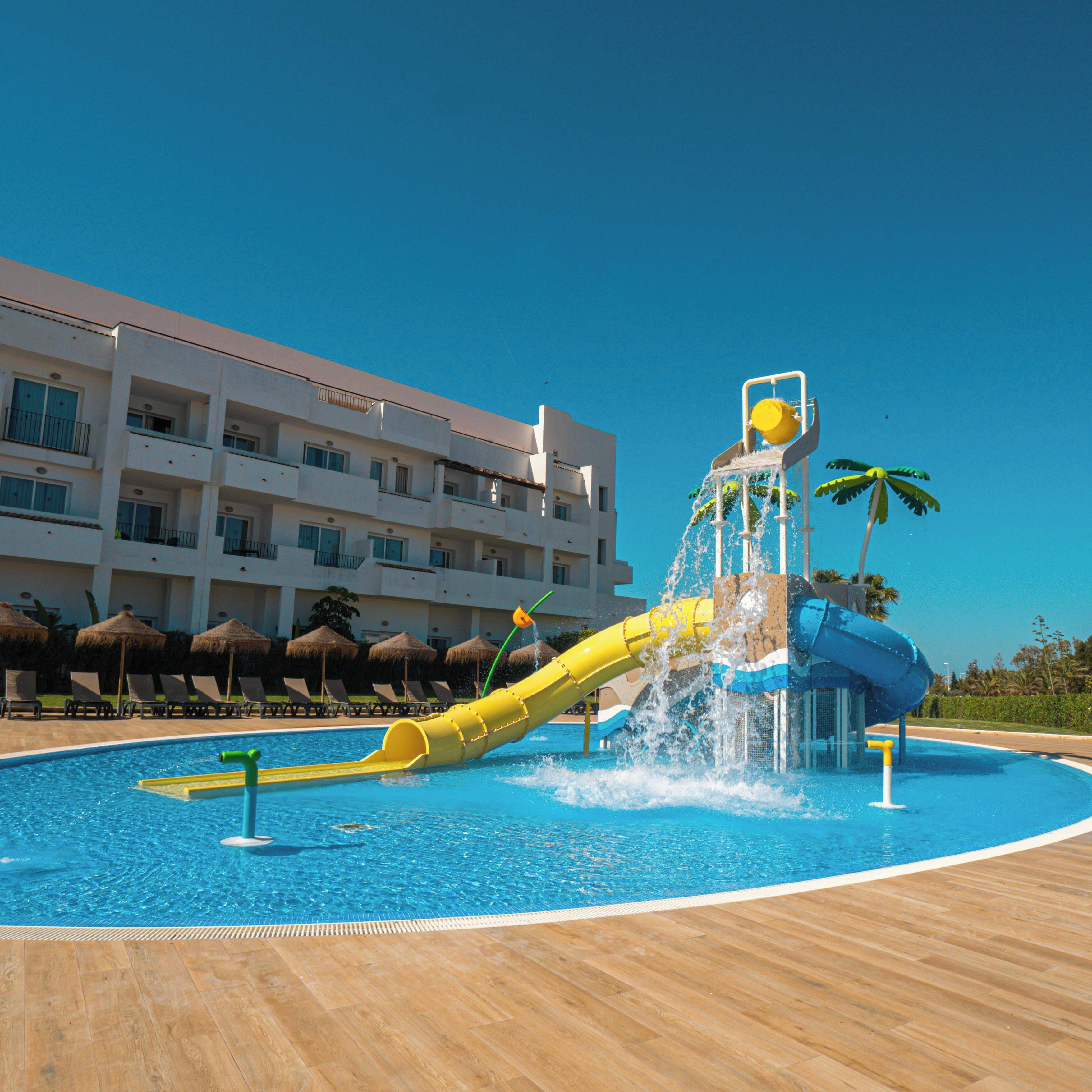 Une piscine avec un toboggan, des chaises longues et un bâtiment d’hôtel en arrière-plan.