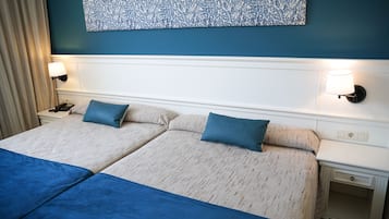 A hotel room with two beds, each with a blue blanket and pillow, a white headboard, and a blue floral artwork above the beds.