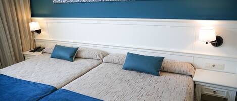 Habitación de hotel con dos camas, cada una con una manta y almohada azules, cabeceros blancos y una obra de arte floral azul sobre las camas.
