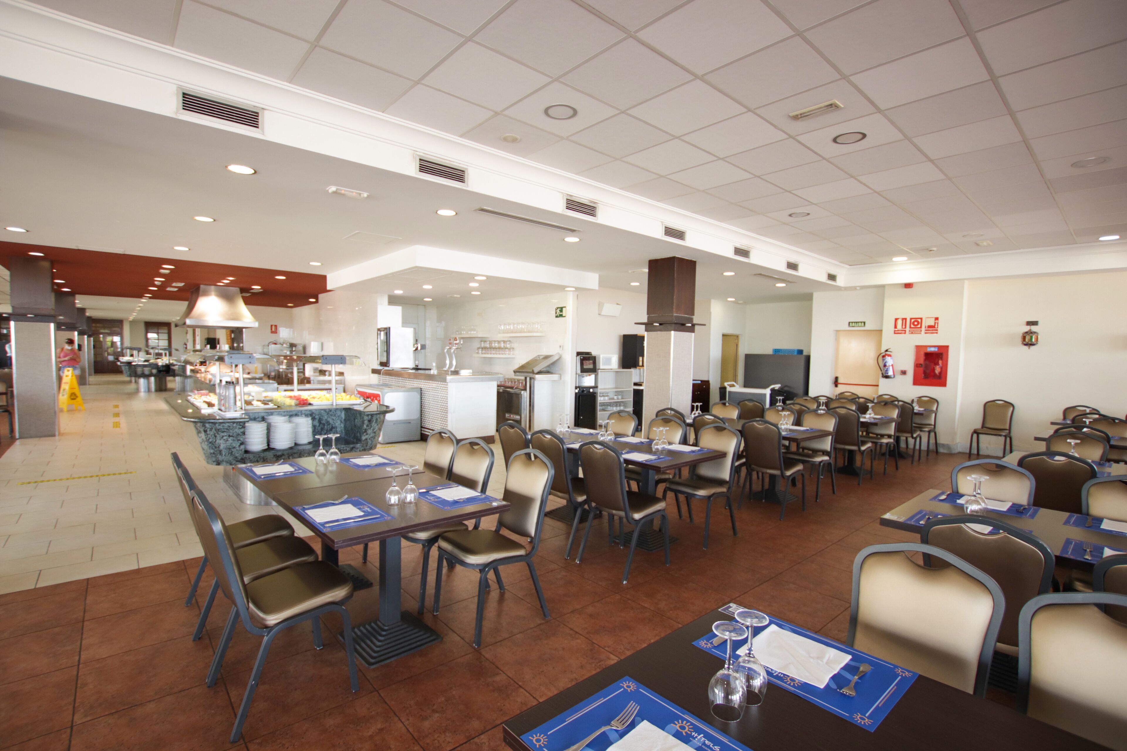 A spacious restaurant with multiple dining tables and chairs, a kitchen area in the background, and a clean, modern interior.