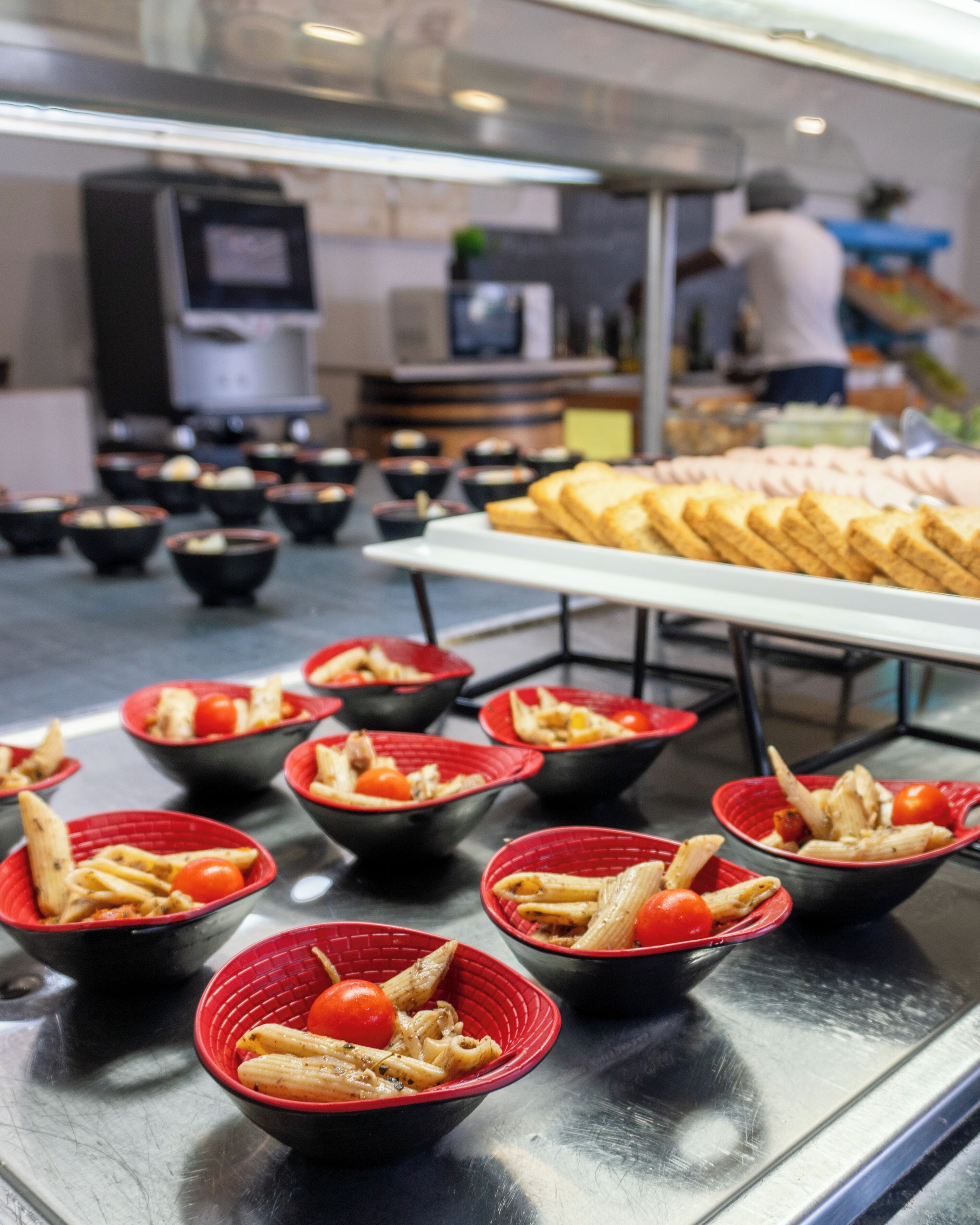 A buffet with pasta dishes in red bowls and a variety of bread on a long table.