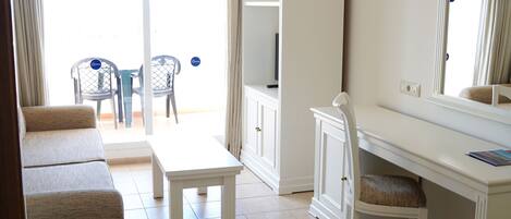 Ein Zimmer mit einem weißen Sofa, einem kleinen Tisch, einem weißen Schreibtisch mit Stuhl, einem großen Spiegel und Blick durch eine Glastür nach draußen.