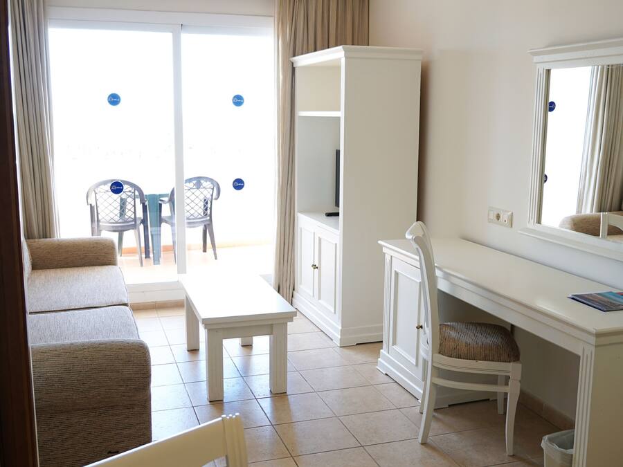 Quarto com sofá branco, mesa pequena, escrivaninha branca com cadeira, espelho grande e vista para o exterior por uma porta de vidro.