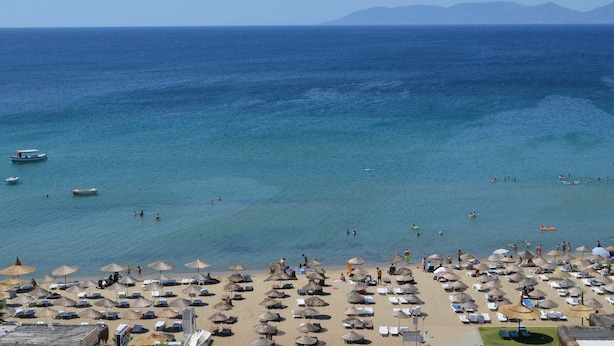 A beach with umbrellas and lounge chairs, a boat in the water, and mountains in the distance. - Grand Milano Hotel (Ayvalik)