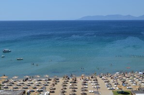 A beach with umbrellas and lounge chairs, a boat in the water, and mountains in the distance. - Grand Milano Hotel (Ayvalik)