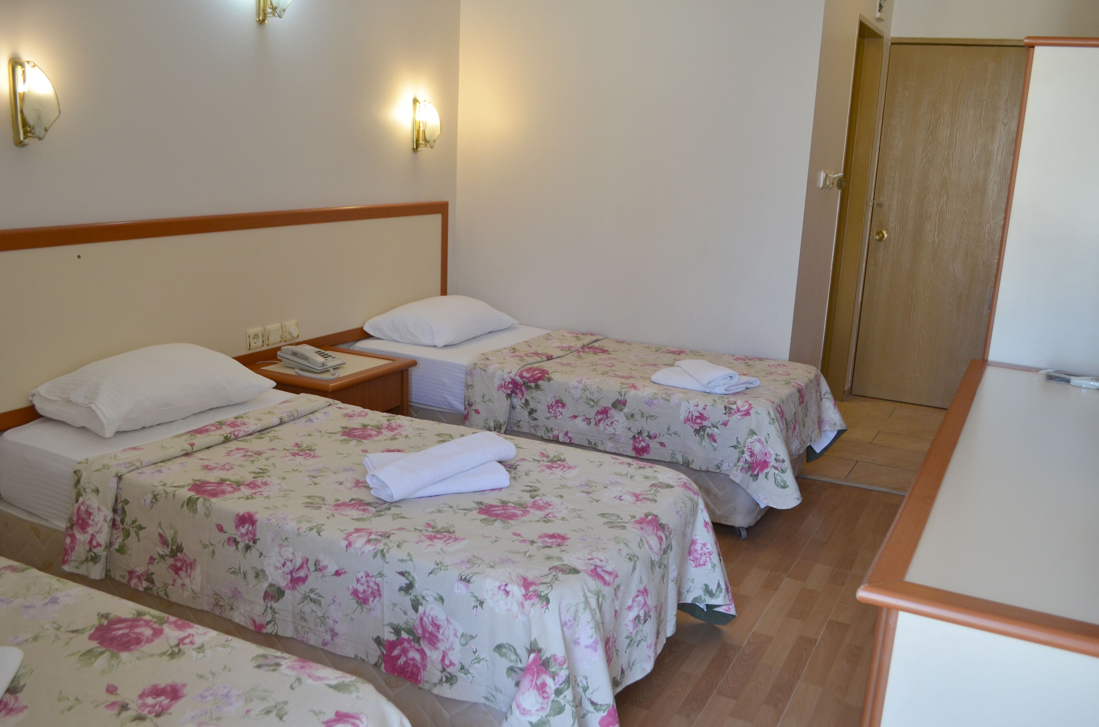 Hotellihuoneessa on kolme erillistä sänkyä, joissa kaikissa on kukallinen päiväpeite ja valkoiset tyynyt. Sängyn vieressä on yöpöydät, joissa on lamput, ja sängyn päädyssä on puinen koristepaneeli. Taustalla näkyy puinen ovi.