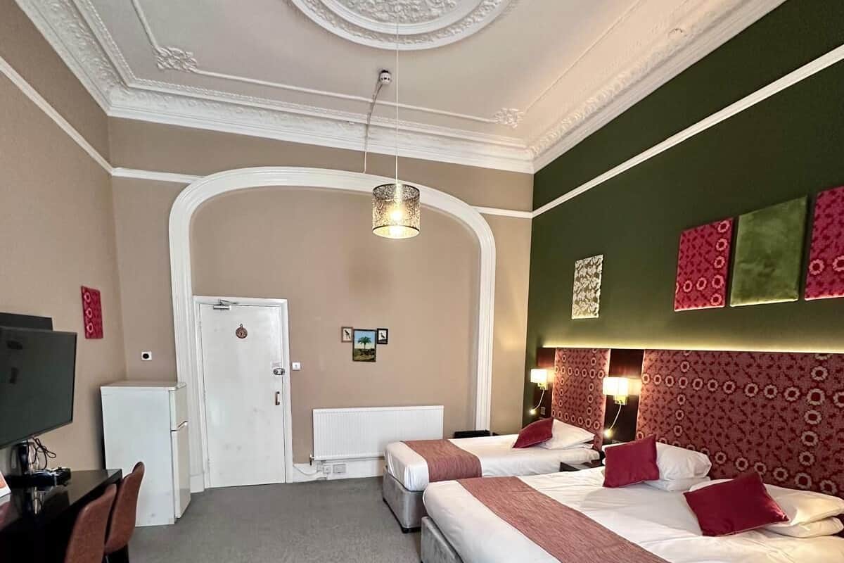 A hotel room with two beds, a desk with chairs, a TV, and a decorative ceiling.