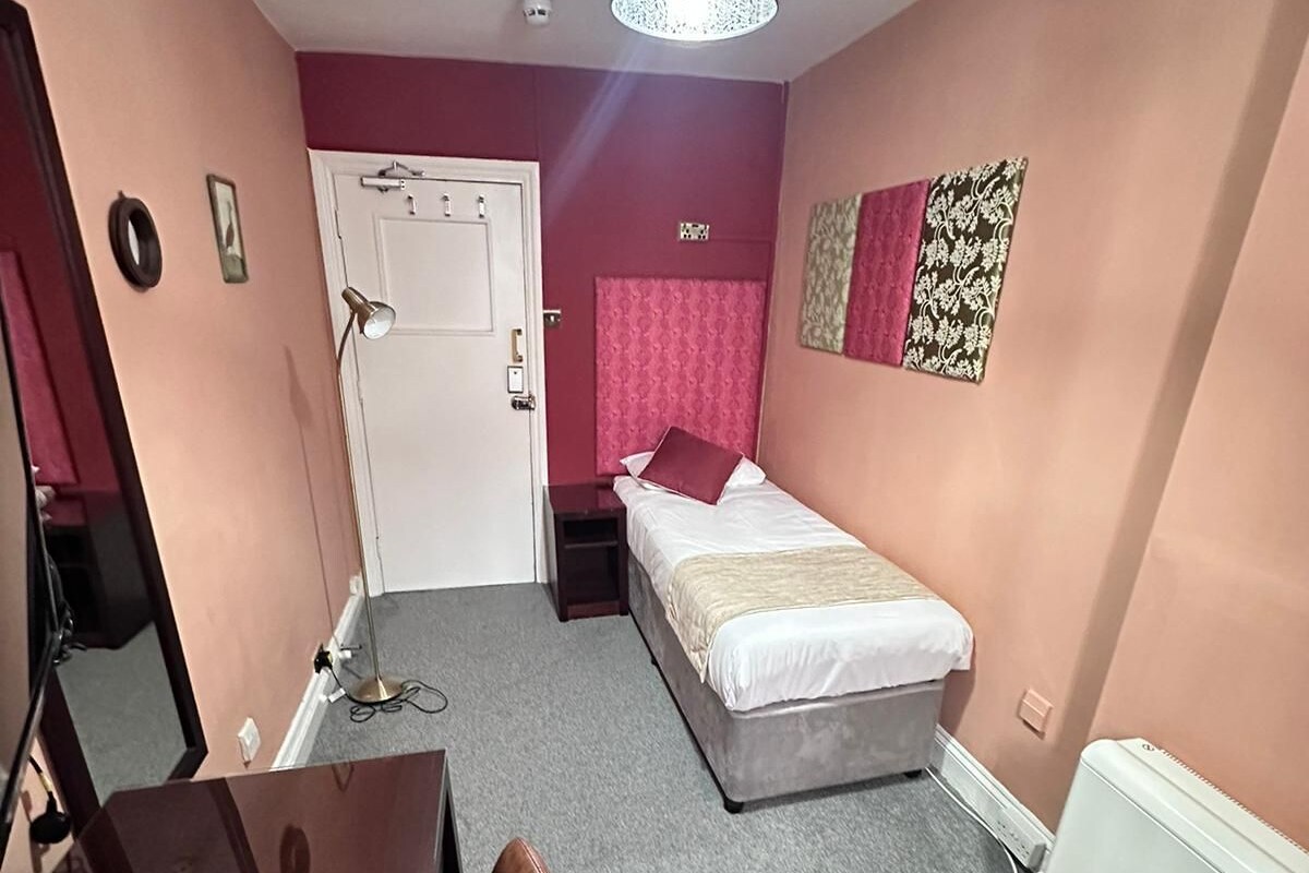 A bedroom with a bed, a desk, a chair, a desk lamp, a radiator, and a ceiling light fixture.