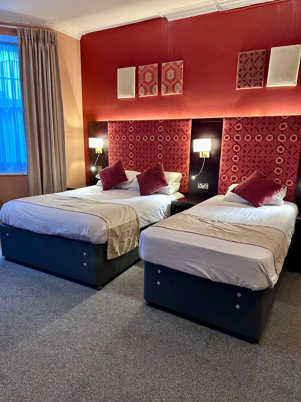 A hotel room with two beds, red patterned headboards, and a window with curtains.