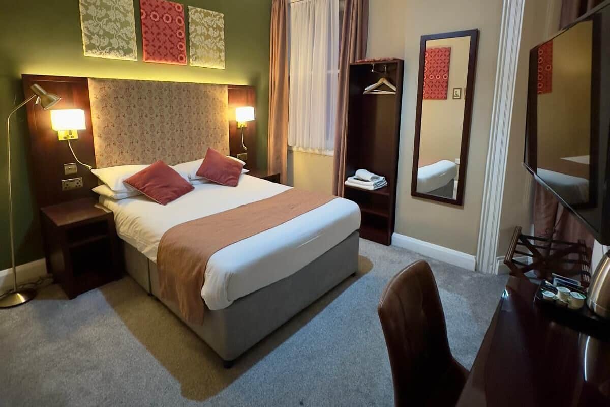 A hotel room with a large bed, bedside lamps, a desk with a chair, a mirror, and artwork on the walls.