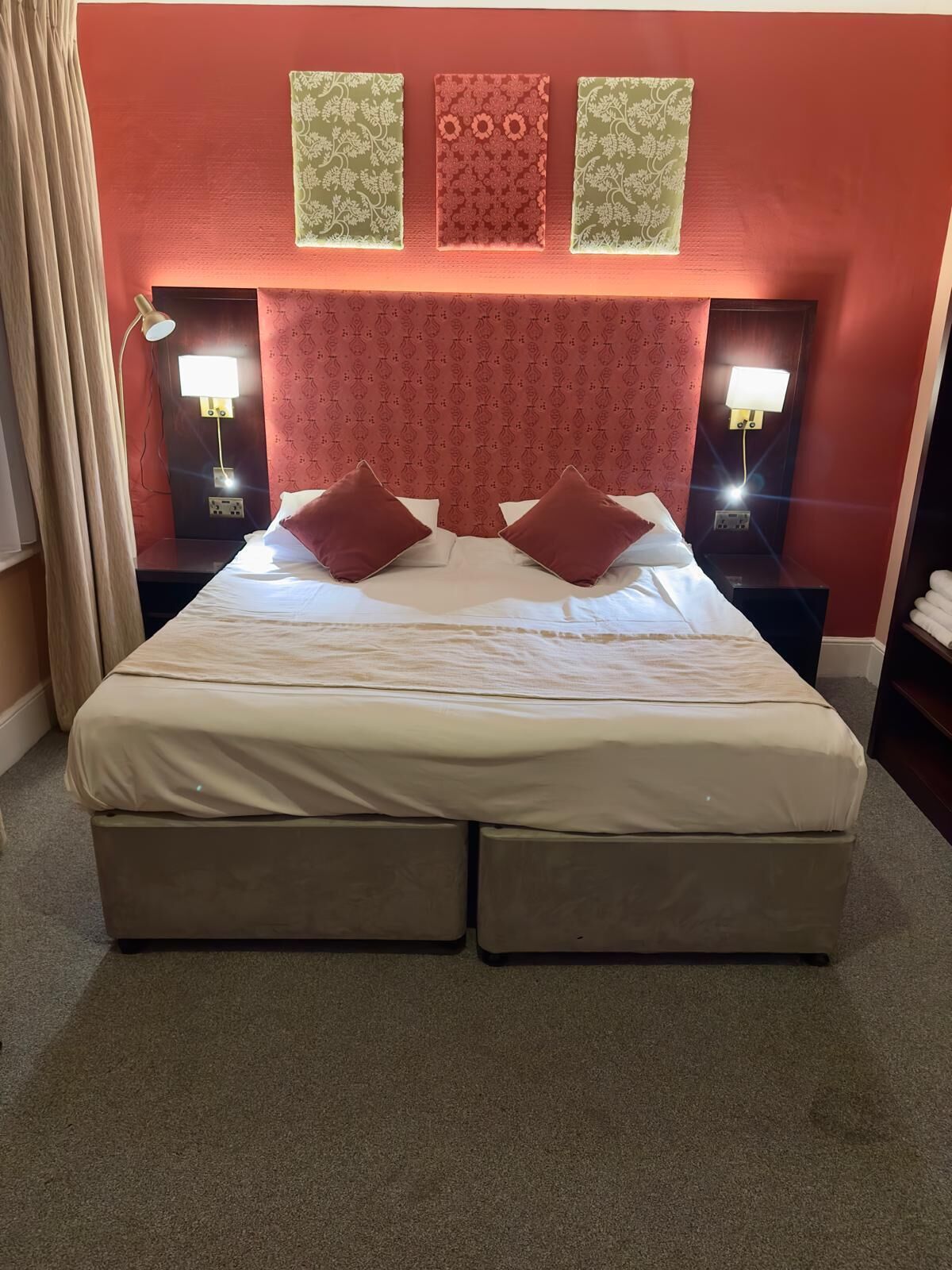 A hotel room with a large bed, two bedside tables with lamps, a headboard with patterned wallpaper, and a curtain on the left.