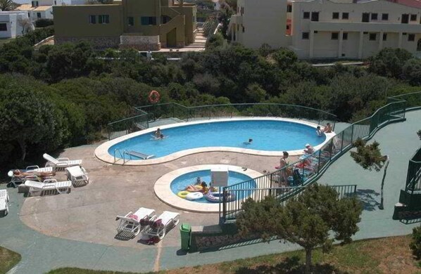 Relax by the pool surrounded by lush greenery and adjacent buildings, featuring a curved design with a central section and a smaller circular area for kids.