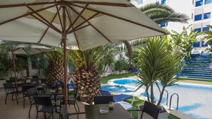 An outdoor seating area with tables and chairs under a large umbrella, overlooking a swimming pool and lush greenery. - Jaime I Hotel Peñiscola (Peniscola)