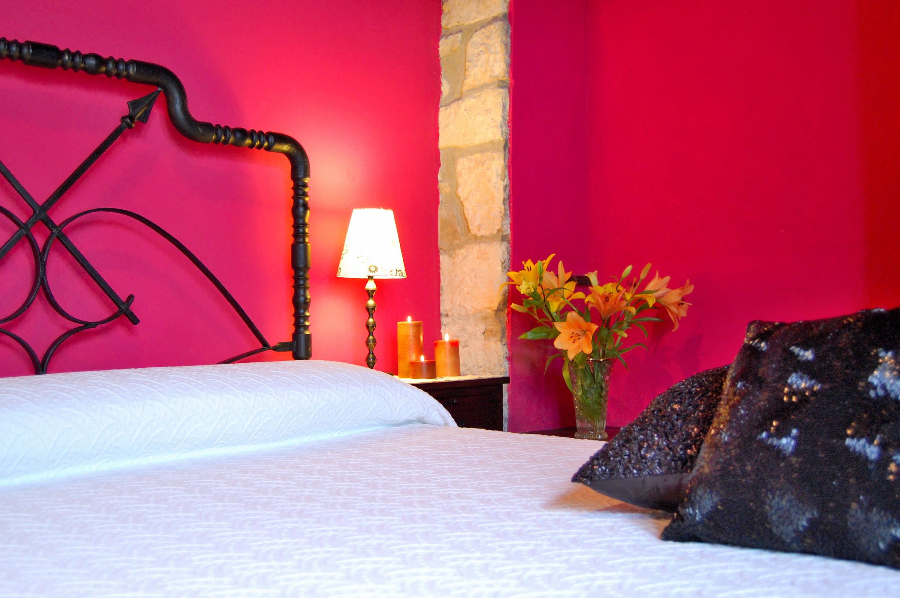 A bed with a white bedspread and black headboard, a bedside table with a lamp, a vase with flowers, and a stone wall.