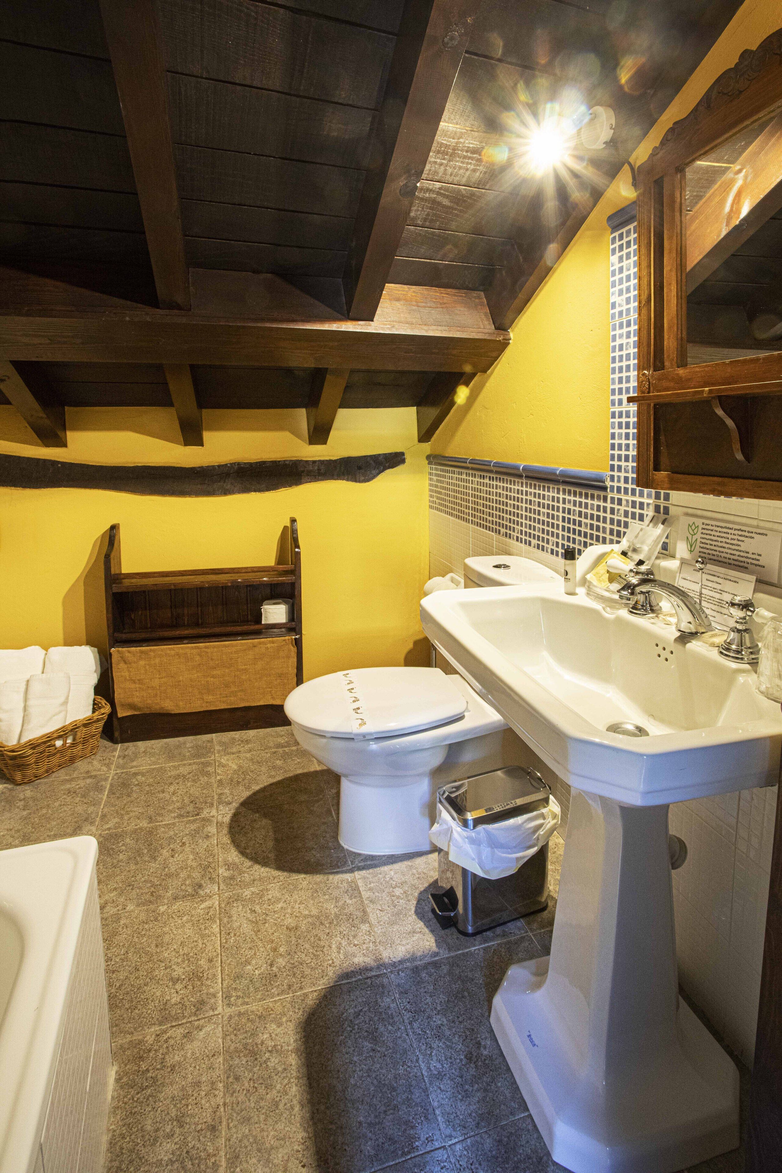 Ein Badezimmer mit Toilette, Waschbecken und Badewanne. Ein Holzregal zeigt Handtücher und einen Mülleimer.