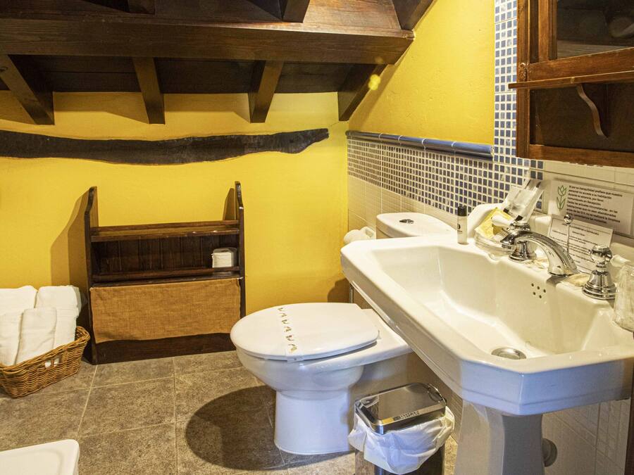 Un bagno con un WC bianco, un lavandino e una vasca. Si vede uno scaffale in legno con asciugamani e un cestino.