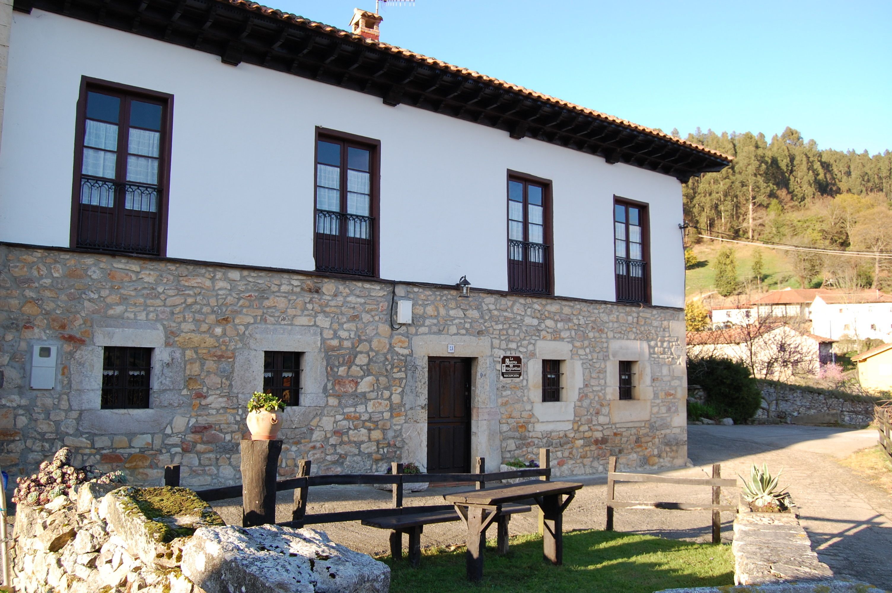 Un edificio de piedra con fachada blanca, varias ventanas con contraventanas oscuras y un área de entrada empavedada.