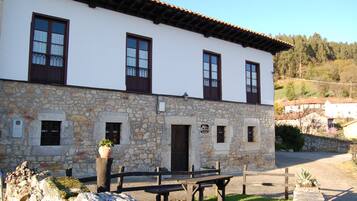 Un edificio de piedra con fachada blanca, varias ventanas con contraventanas oscuras y un área de entrada empavedada.