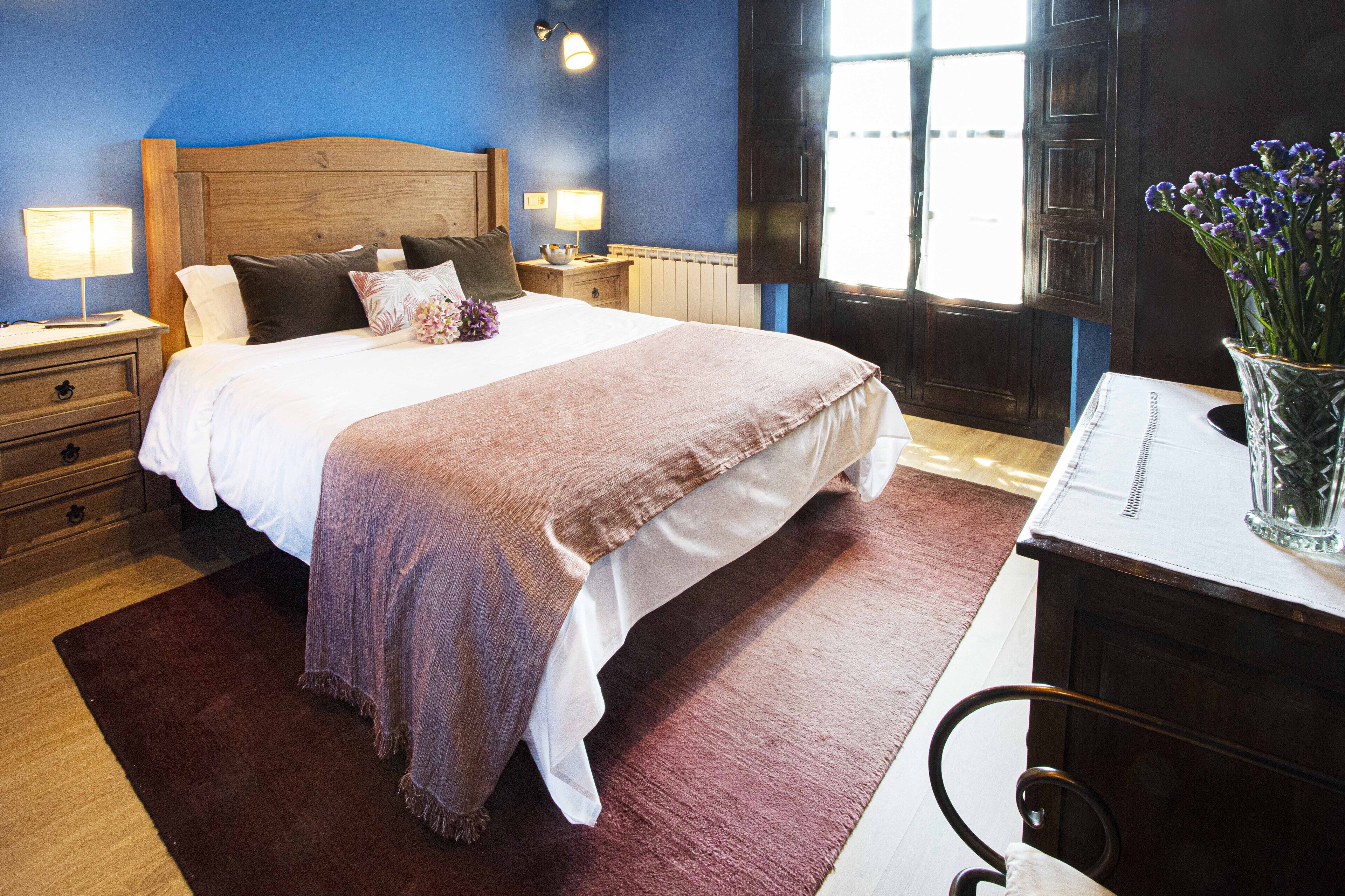 Un dormitorio con una cama grande, cabecera de madera, mesitas de noche con lámparas, una mesita auxiliar, un aparador y una ventana con cortinas.