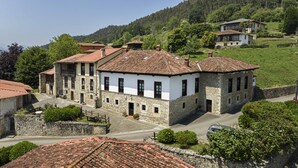 Un edificio de piedra tradicional con tejado de tejas, rodeado de vegetación y otros edificios.