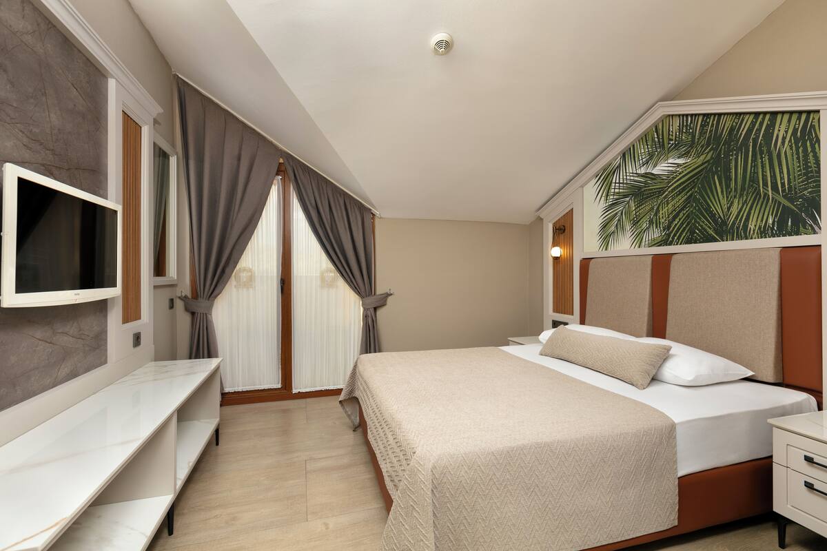 A modern hotel room with a large bed, a TV mounted on the wall, and a window with curtains.