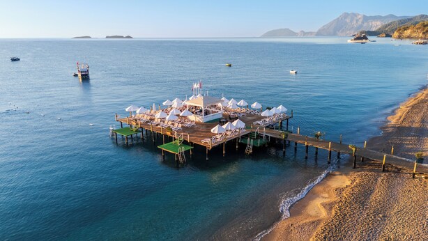 A pier with lounge chairs and umbrellas, extending into a calm sea with boats and distant mountains. - Club Hotel Phaselis Rose - All Inclusive (Kemer)