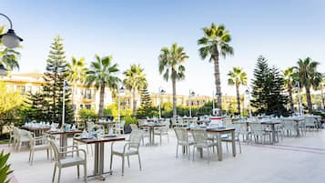 An outdoor dining area with tables and chairs, surrounded by palm trees and buildings.