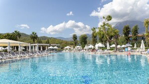 A large pool with multiple sun loungers and umbrellas, surrounded by palm trees and mountains in the background. - Club Hotel Phaselis Rose - All Inclusive (Kemer)