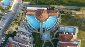 An aerial view of a resort with a large central pool, multiple smaller pools, and a circular arrangement of sun loungers and umbrellas. - Exotica Hotel (Zakynthos)