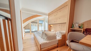 A bedroom with a wooden bed, a wooden desk, and a chair. There is a window with a view of the outside. - Hotel Ischgl (Ischgl)