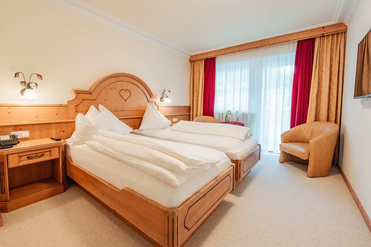 A hotel room with two beds, wooden headboards, a nightstand, a chair, and a large window with red curtains.