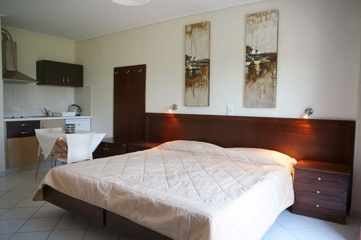 A bedroom with a large bed, a bedside table, a nightstand, a desk with a chair, and a kitchenette in the background.