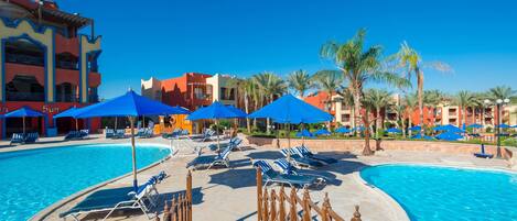 Zona junto a la piscina con sillones de descanso y sombrillas, una valla de madera y un cielo azul despejado.