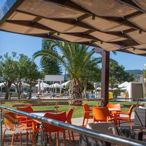 An outdoor seating area with orange chairs and tables, a palm tree, and a pool in the background. - Ialyssos Bay (Rhodes)