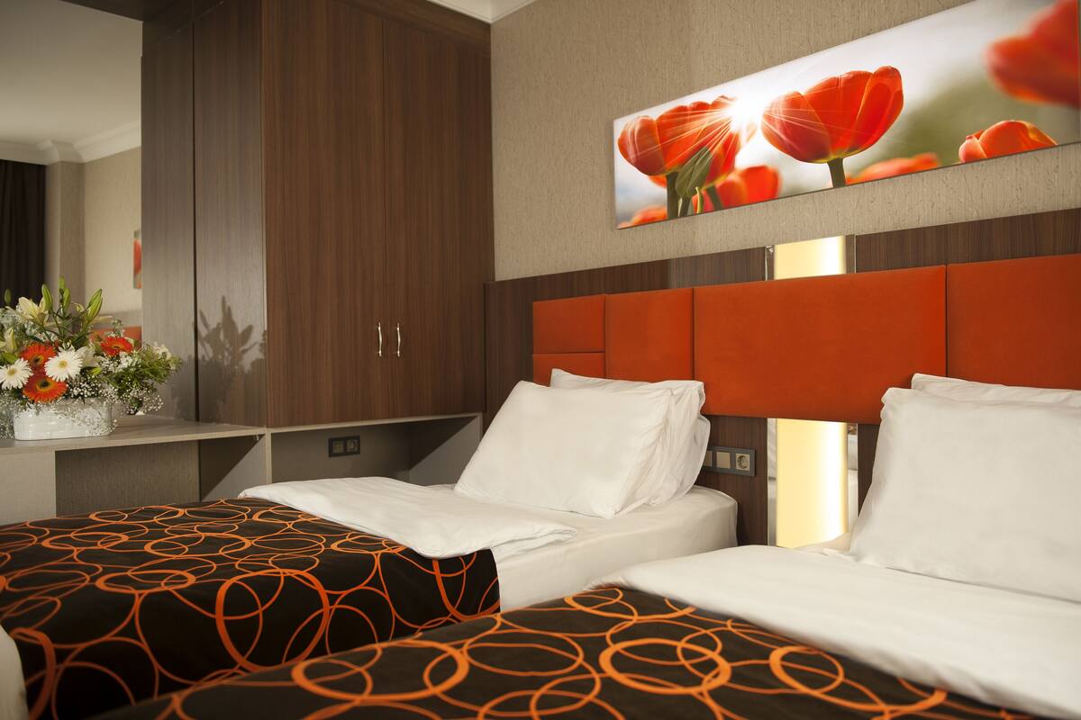 A hotel room with two beds, a wooden wardrobe, a wall-mounted TV, and a floral arrangement on the nightstand.