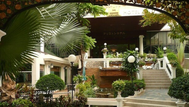 A hotel courtyard with a central fountain, surrounded by potted plants and a stone pathway leading to the entrance. - The Ariston (Bangkok)