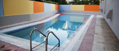 Una piscina con una escalera metálica, rodeada por un suelo de baldosas y un muro colorido.