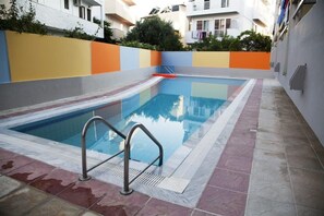Una piscina con una escalera metálica, rodeada por un suelo de baldosas y un muro colorido.