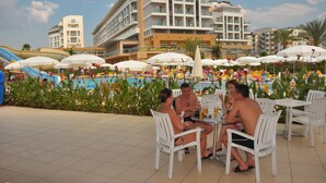 Ein Poolbereich mit weißen Stühlen und Sonnenschirmen, einem Schwimmbecken und einem Gebäude mit Balkonen im Hintergrund.