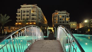 Ein Nachtblick auf ein Hotel mit Poolbereich, einschließlich einer Brücke mit Metallgeländern zum Pool.