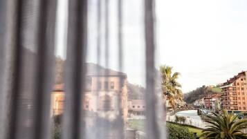 A view through a window with curtains, featuring a river, buildings, and a palm tree.