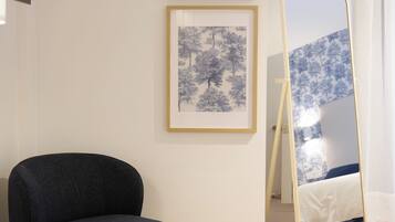 A room with a dark blue armchair, a small round table, two framed blue tree pictures on the wall, and a large mirror reflecting a bed and window.